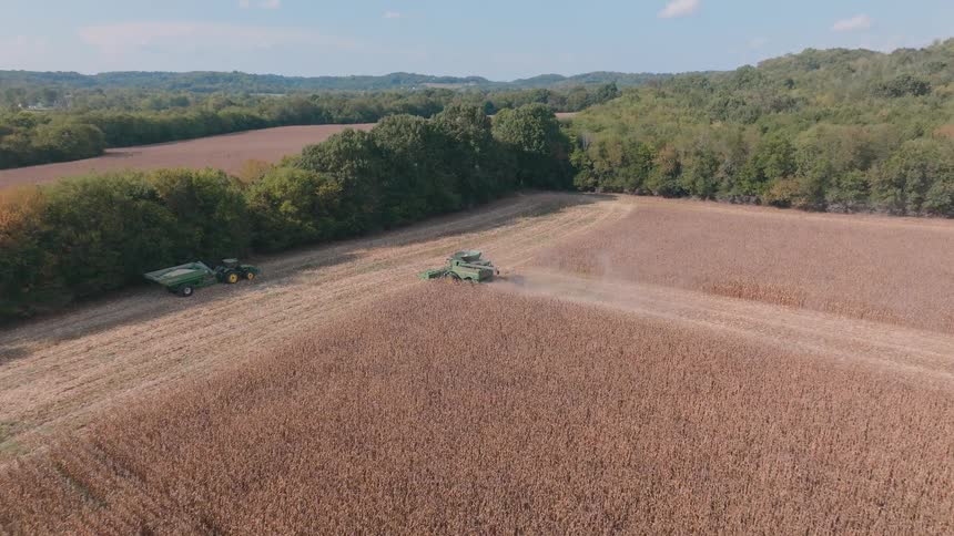 High aerial view of a green combine harvester moving through a vast, dry cornfield during the autumn harvest, with a tractor and grain cart nearby