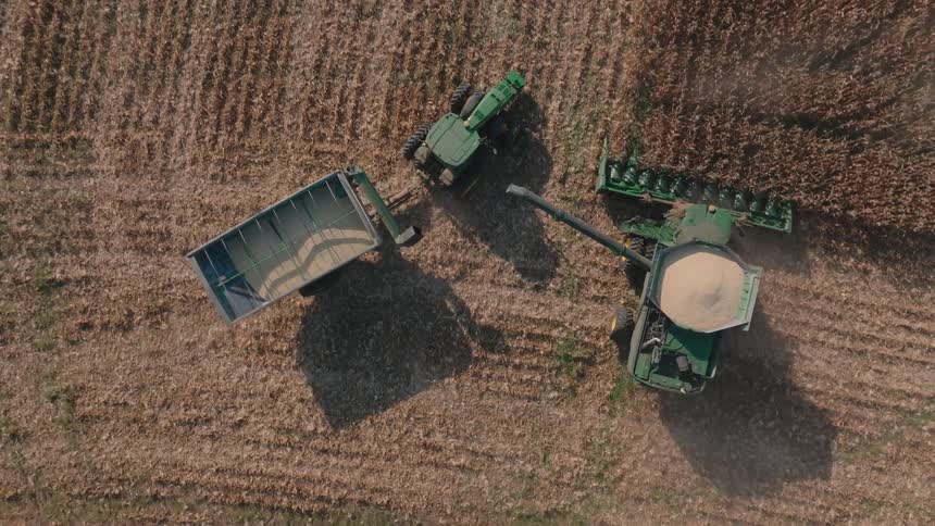An aerial top-down view captures a combine harvester unloading harvested corn into a tractor-trailer while continuing to work in a vast farm field