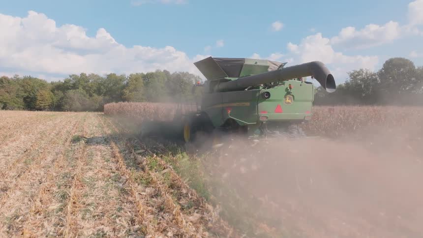 A combine harvester kicks up clouds of dust and chaff while working through a cornfield in slow motion on a sunny day