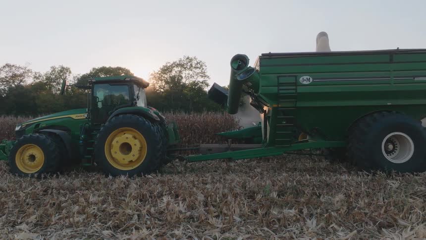 A combine offloads harvested corn into a grain cart pulled by a tractor in a field at sunset