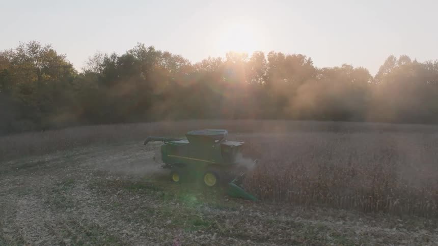 A combine harvester kicks up dust while harvesting crops in a field during a hazy golden sunset