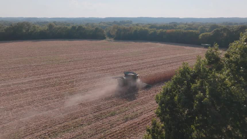An aerial view reveals a combine harvester moving through a vast field, kicking up dust as it gathers crops on a sunny day