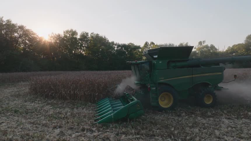 An aerial view captures a combine harvester moving through a cornfield at sunset, gathering the crops in slow motion