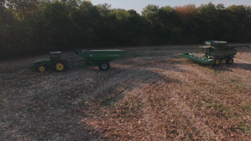 An aerial view shows a combine harvester offloading its grain into a waiting tractor and grain cart in a rural field