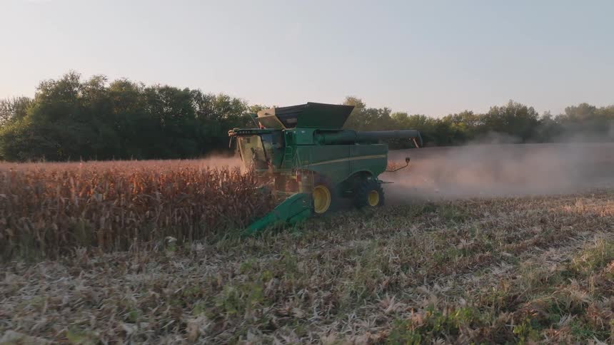 A combine harvester moves through a cornfield in slow motion, kicking up dust as it harvests the crop during a beautiful sunset