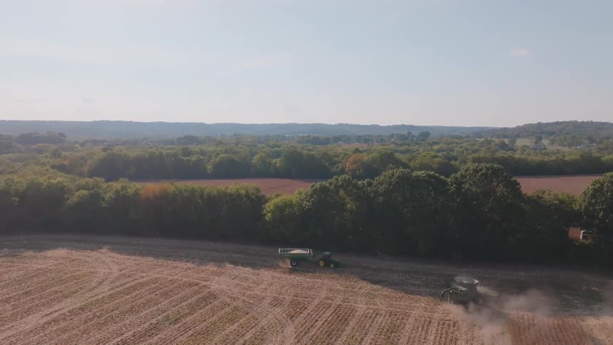 A combine harvester and tractor work in tandem to harvest crops in a vast field, as seen from a wide aerial perspective