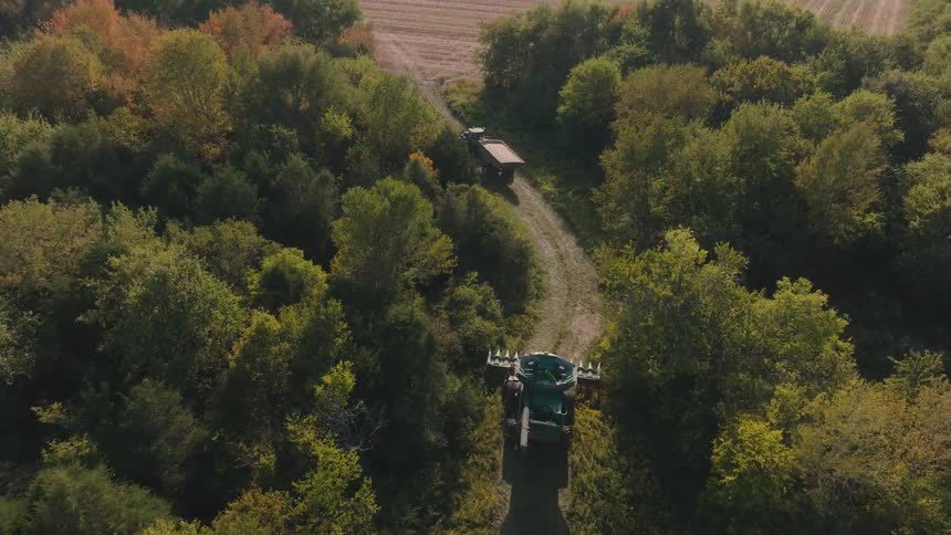 An aerial shot slowly rises above a combine harvester on a wooded path, revealing a large harvested farm field and the surrounding forest