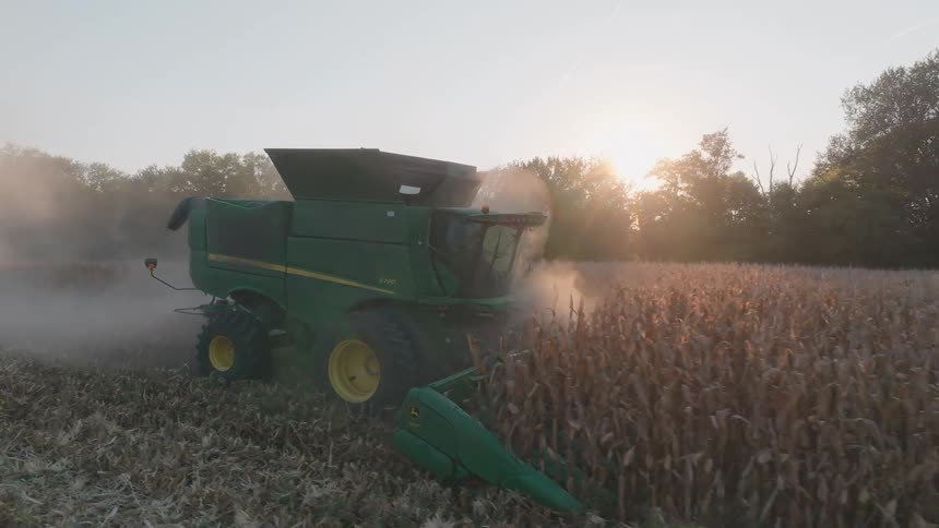 A green combine harvester cuts through a dusty field of corn as the sun sets, creating a hazy, golden glow