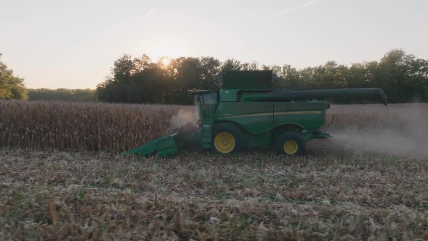 A combine harvester drives through a vast cornfield at sunset, kicking up dust as it gathers the autumn crop