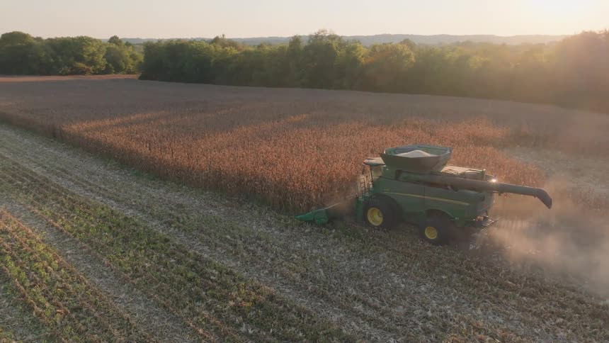 An aerial view shows a combine harvester kicking up dust as it works through a cornfield during a beautiful golden hour sunset