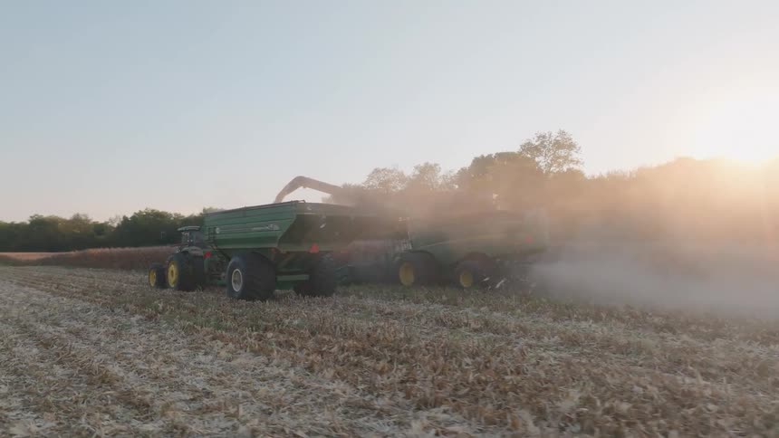 A combine harvester offloads harvested corn into a grain cart pulled by a tractor in a dusty field during a beautiful sunset