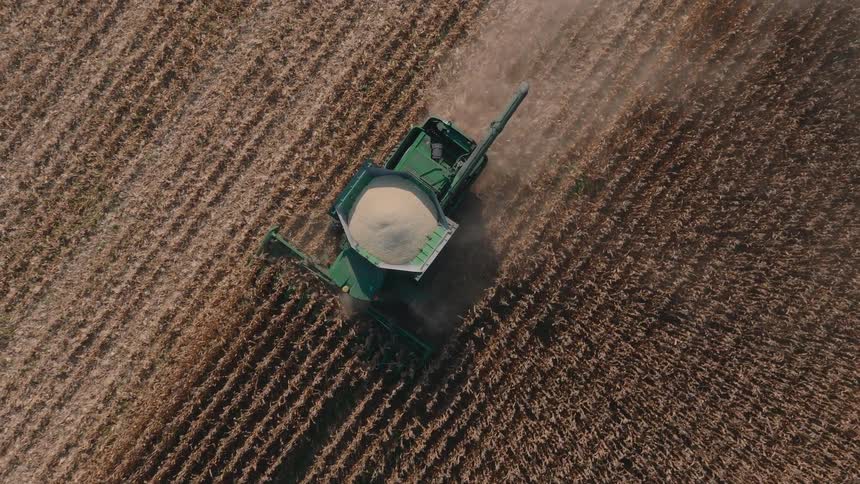 An aerial top-down view shows a green combine harvester moving through a dry cornfield, collecting the crop during the autumn harvest