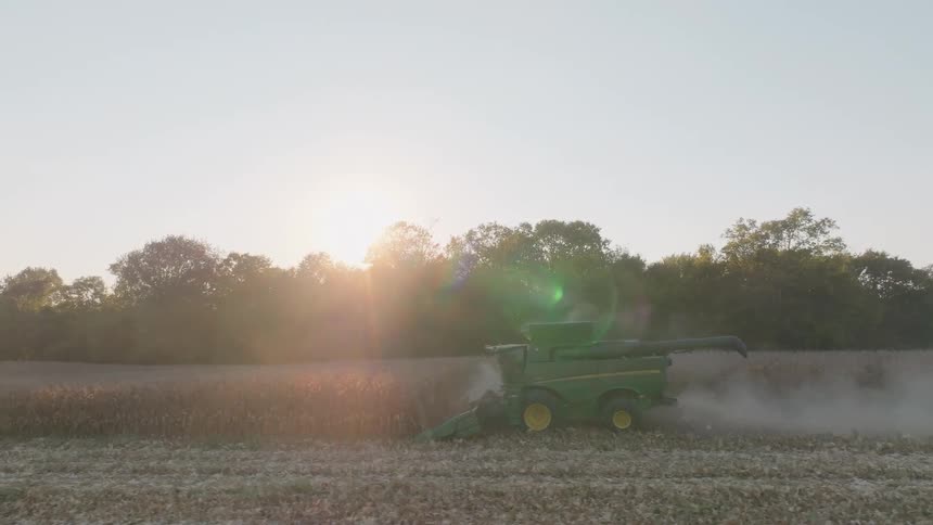 In slow motion, a combine harvester kicks up dust while working in a rural crop field as the golden sun sets behind a line of trees