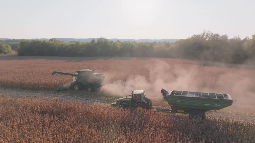 An aerial view of a combine harvester offloading its payload of corn into a tractor's grain cart during a dusty harvest