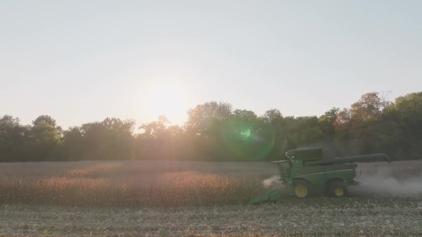 A slow-motion wide shot captures a combine harvester kicking up dust as it harvests a vast crop field during a beautiful golden hour sunset
