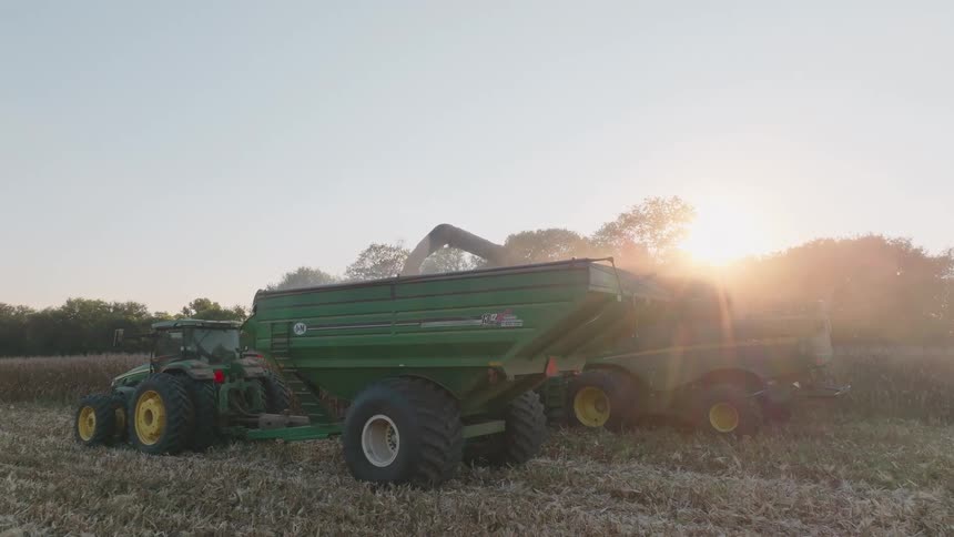 A combine harvester transfers its yield into a tractor-pulled grain cart as the golden sun sets over a dusty farm field