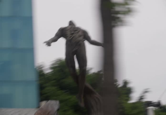 A blurry, shaky, handheld view of a muscular male statue with arms outstretched in an urban setting