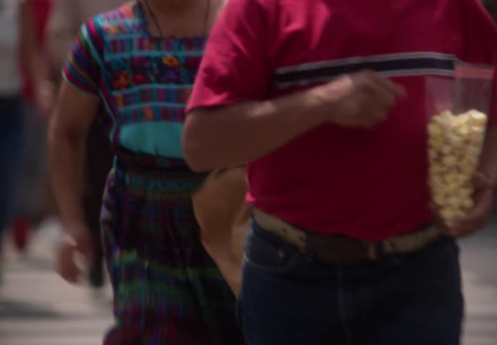 A man in a red shirt holds a bag of popcorn while walking with a crowd across a city crosswalk in slow motion