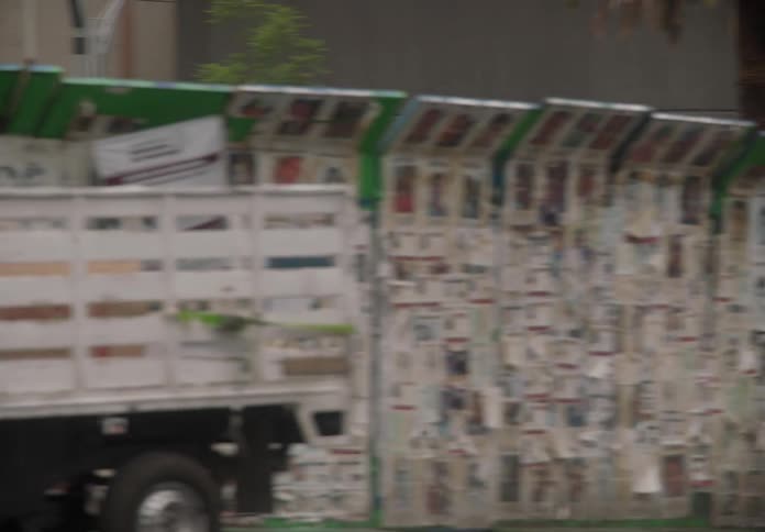 A slow-motion pan of a Mexico City street reveals a police truck, a double-decker bus, and a long wall covered in missing persons posters