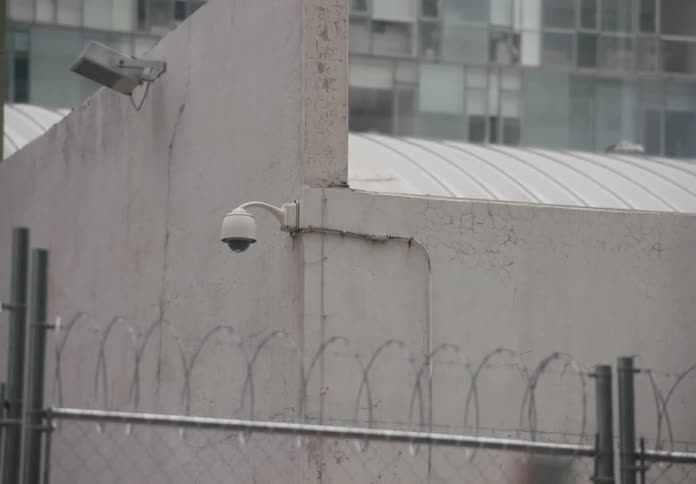A fast whip pan shot transitions from a blurry city street to a security camera on a concrete wall with a barbed wire fence