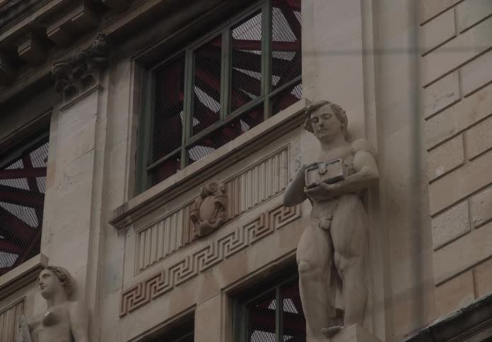 Slow zoom towards an ornate stone statue of a man holding a savings box on a classical building facade
