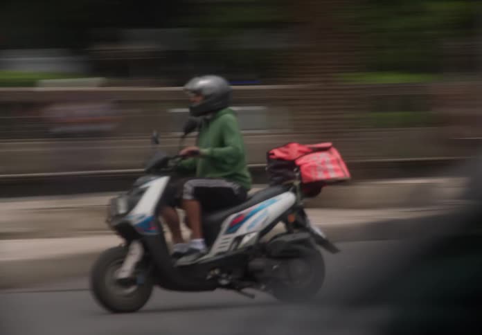 A delivery driver on a scooter swiftly navigates through blurry city traffic