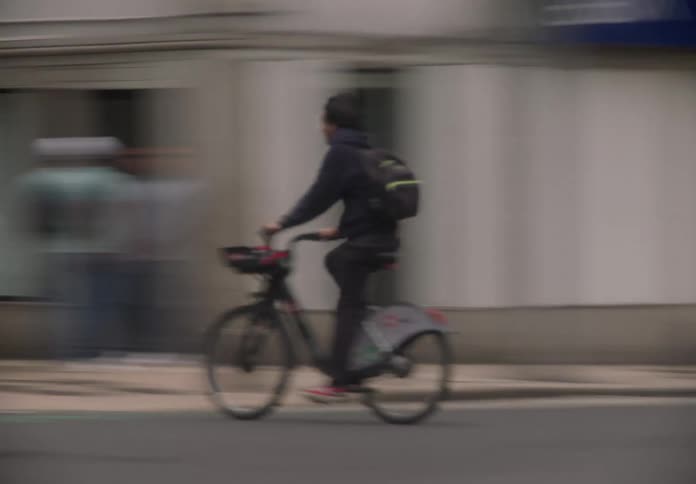 Side view pan shot captures a young man with a backpack riding a bicycle through a bustling city street with a motion blur effect