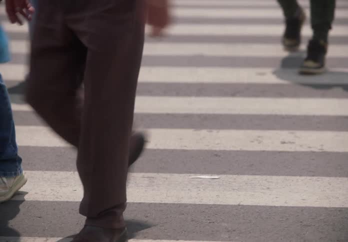A low-angle slow motion shot captures the legs and feet of a diverse crowd of pedestrians walking across a city crosswalk on a sunny day