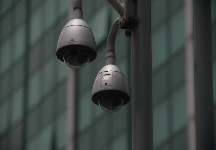 A quick whip pan reveals a close-up of two dome security cameras mounted on a pole in front of a modern glass building