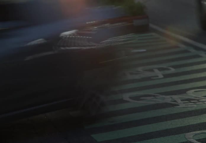 A blurry car with bright headlights drives over a green striped bicycle lane on a city street at night