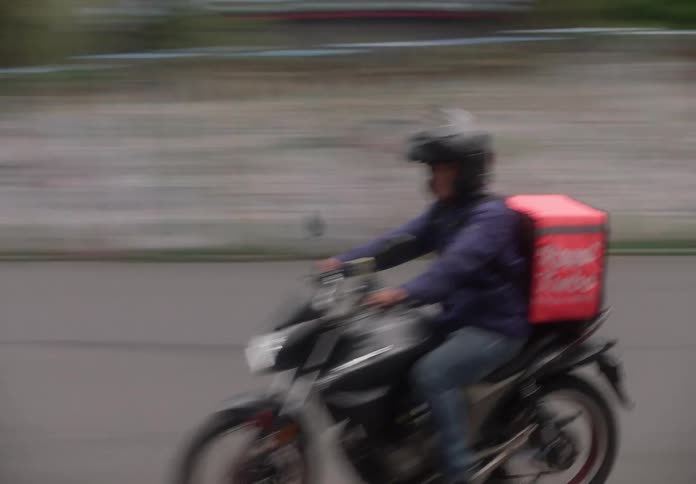Tracking shot follows a Rappi Turbo food delivery driver speeding on a motorcycle through busy city streets