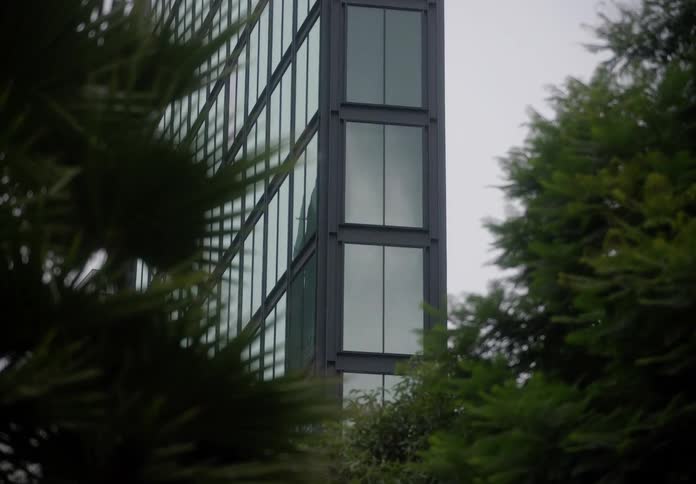 A modern glass office building is framed by lush green trees on a cloudy, overcast day