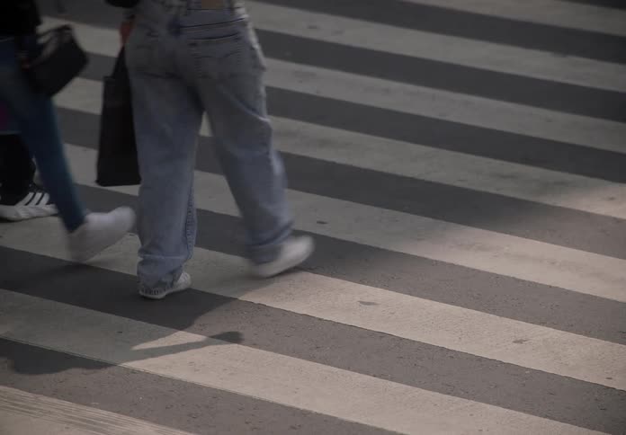 A close-up, high-angle view of the legs and feet of various pedestrians walking across a striped crosswalk on a city street