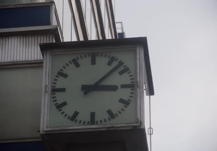 Slow zoom out from a large square clock attached to the side of a modern urban building
