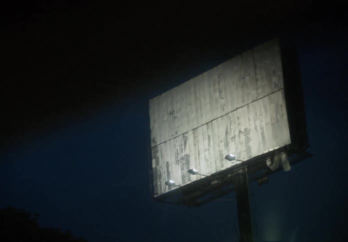 An old, empty billboard is lit against the night sky before being obscured by a passing shadow