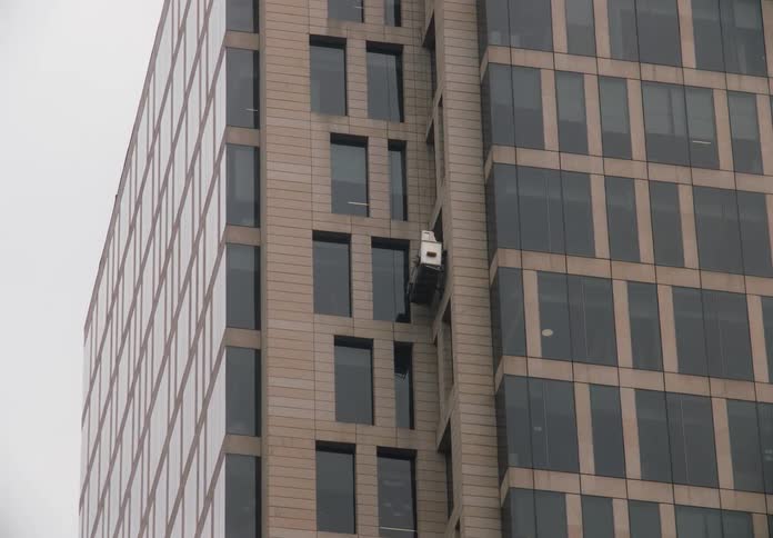 A window washing gondola slowly descends the side of a modern glass skyscraper on an overcast day