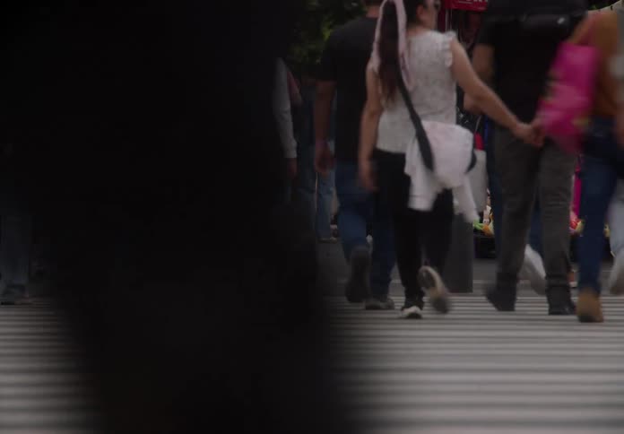 A low-angle time-lapse captures the blurred motion of a diverse crowd's legs and feet as they cross a busy city street