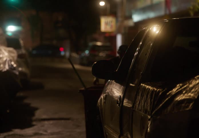 Slowly zooming in on a covered car parked on a dimly lit, gritty city street at night