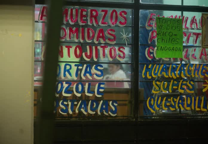 Hand-painted menu signs for traditional Mexican food adorn the window of a small, local eatery at night