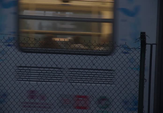 Side view of a colorful commuter train speeding past a chain-link fence at dusk before coming to a stop
