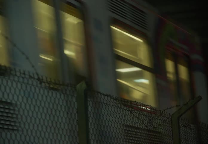 A graffiti-covered metro train speeds through a dark urban tunnel at night, viewed from behind a chain-link fence