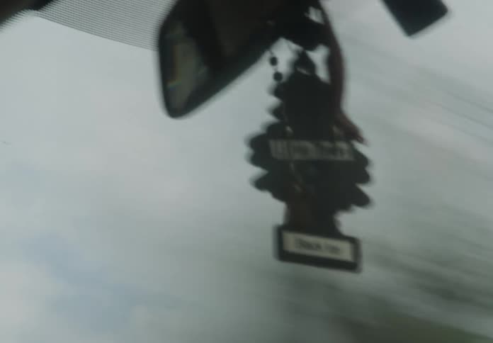 Point-of-view shot from inside a car focuses on a swinging air freshener and rosary against a blurred, moving background