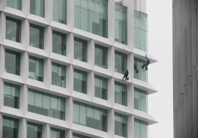 Two window washers rappel down the side of a modern white office building to clean the exterior glass