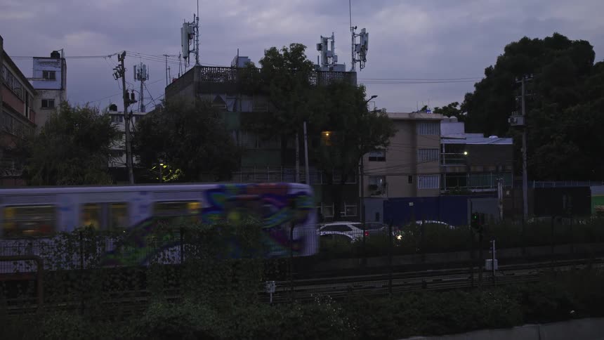 A graffiti-covered subway train travels alongside a busy city street during a cloudy evening