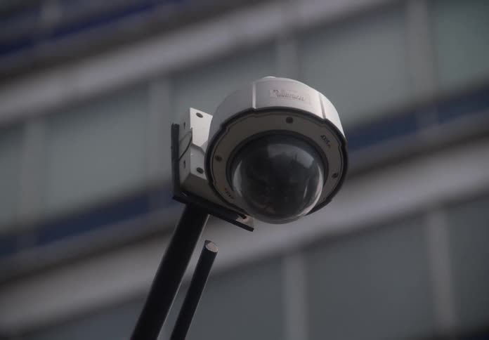 A slow zoom out from a dome-style surveillance camera mounted outside a large office building