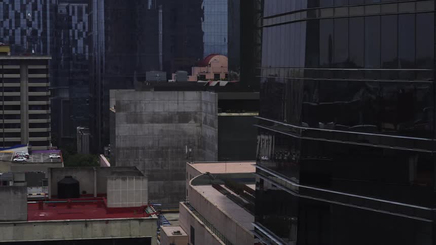 Slow high-angle tracking shot moves past a modern glass skyscraper revealing the diverse urban rooftops of a city on an overcast day