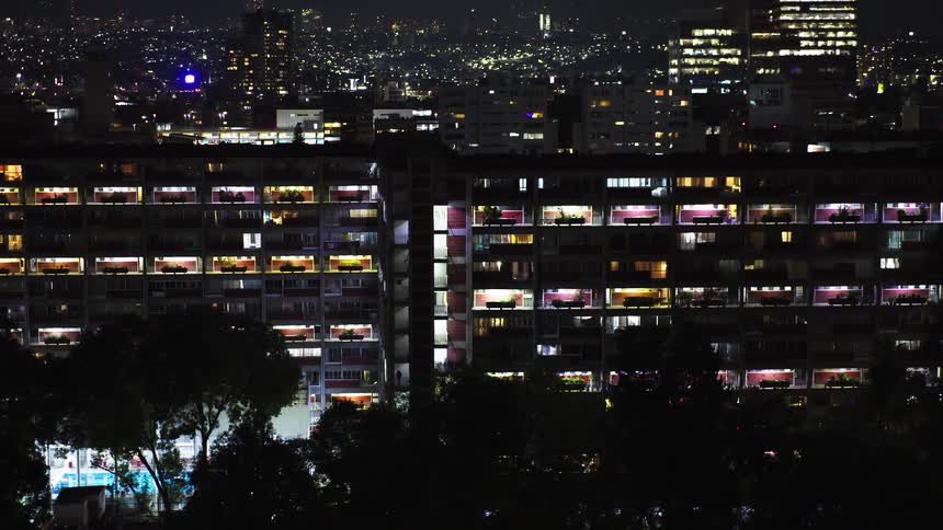 A slow pull-back shot reveals a vast apartment complex illuminated at night, with a brightly lit cityscape in the distance