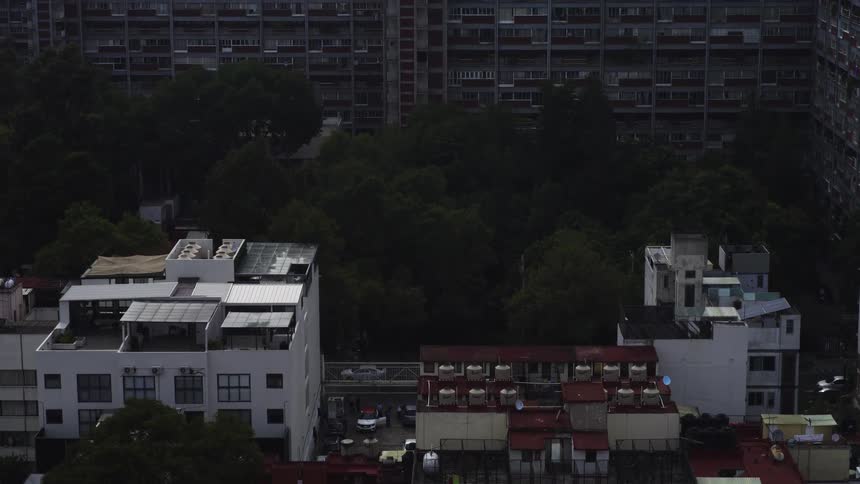 An aerial shot slowly ascends over residential rooftops, revealing a sprawling urban apartment complex behind a dense patch of trees on an overcast day
