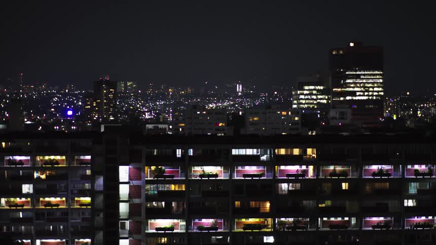Drone shot pulls back from a glittering city skyline at night to reveal a large apartment building with residents in their illuminated homes