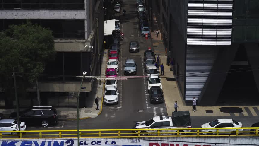 A high-angle zoom-out shot captures daily life on a narrow urban street lined with parked cars and modern office buildings